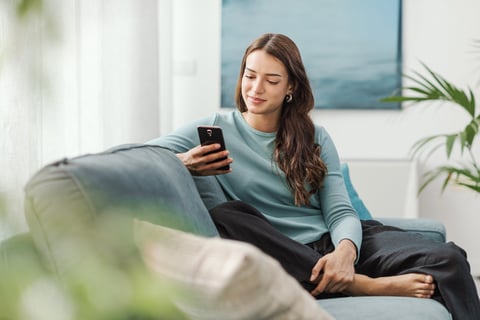 Happy young woman sitting on the couch at home and chatting with her smartphone
