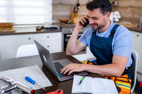 Handyman discussing heating repair details on phone while using laptop at kitchen table.